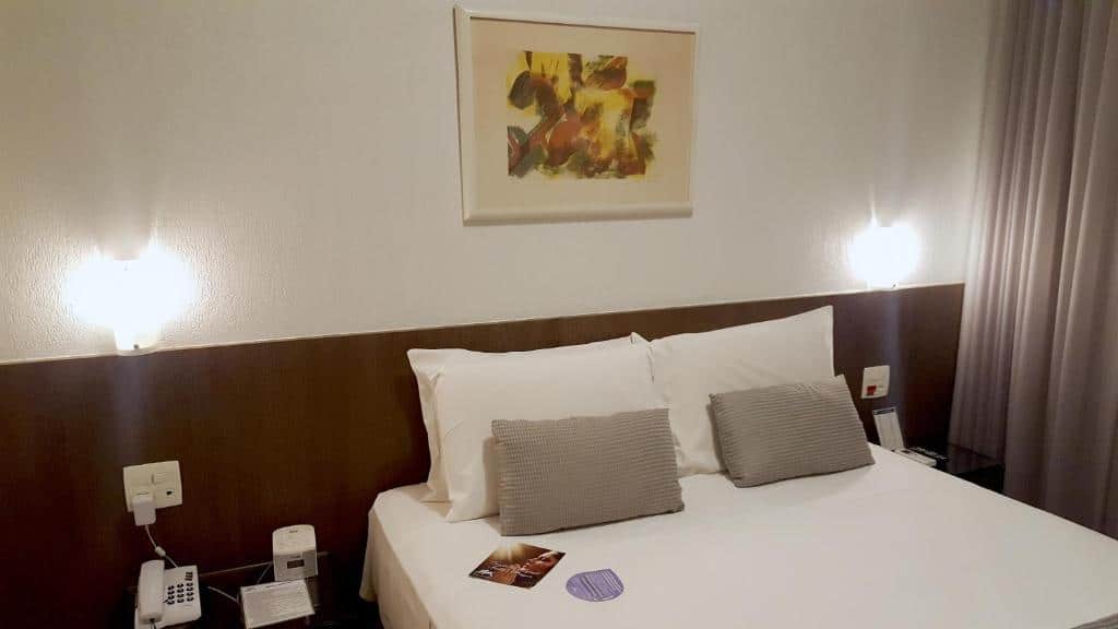 Foto do Condomínio Max Savassi Superior apto 1303. O quarto possui uma cama de casal no centro da parede e uma escrivaninha em seus dois lados. Na direita vemos parte da cortina. Representa o post de airbnb em Belo Horizonte.