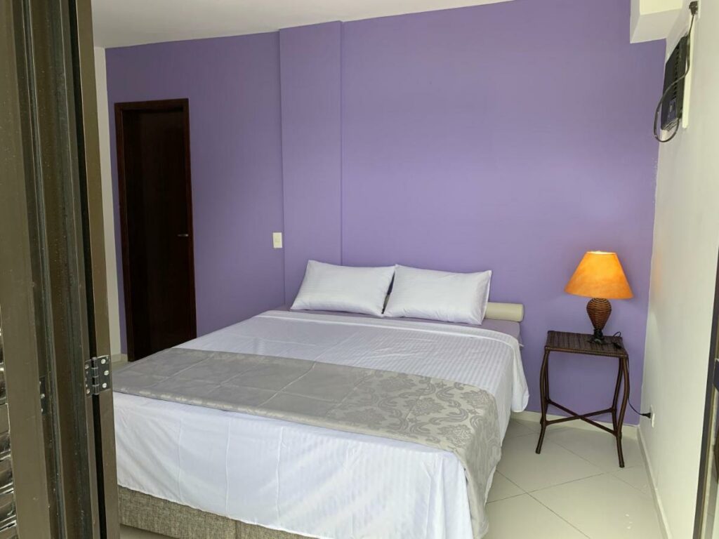 Quarto do Lindo Duplex Pé Na Areia com Praia Privativa, um dos airbnb em Caraguatatuba. Uma cama de casal com jogo de cama e travesseiros tem uma mesinha de cabeceira com abajur ao lado direito e uma porta ao lado esquerdo. A foto foi tirada do lado de fora da porta que leva à sacada.
