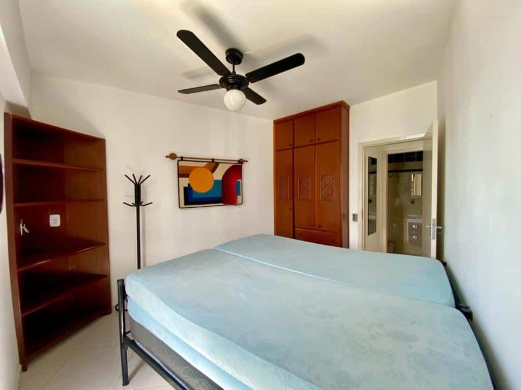 Quarto do APTO DE FRENTE PARA O MAR - CARAGUATATUBA, um dos airbnb em Caraguatatuba. Na parede ao lado direito há duas camas de solteiro encostadas, que encaram uma estante e um cabideiro na parede em frente. Logo acima há um ventilador de teto. Na parede ao lado direito das camas fica uma porta que leva ao banheiro e um guarda-roupa de madeira.