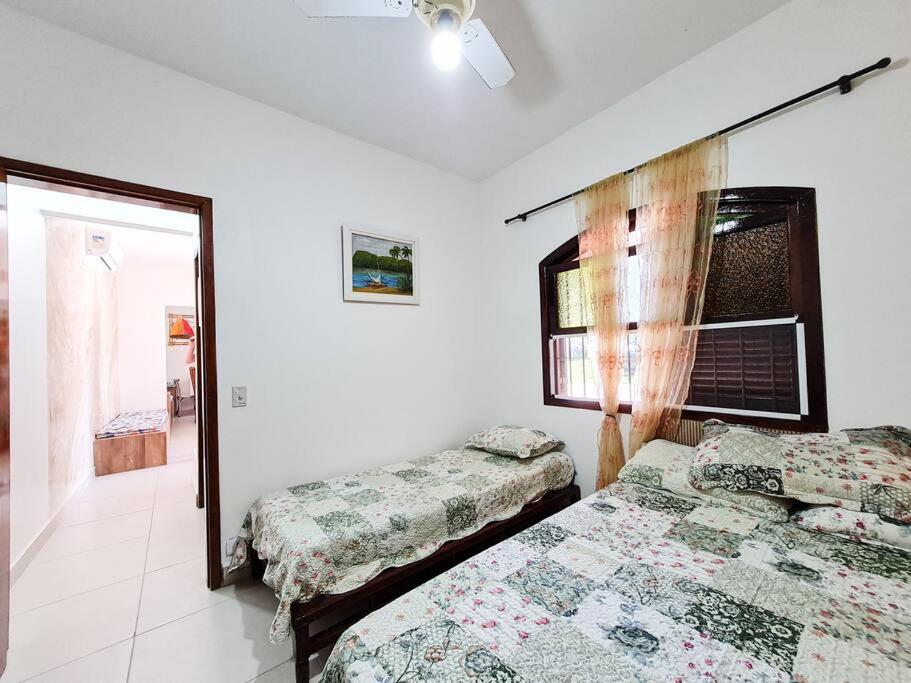 Quarto da Casa vista para montanha Pet Friendly, um dos airbnb em Caraguatatuba. Uma cama de solteiro está encostada na parede ao lado esquerdo, e a porta do quarto está aos seus pés. Uma janela fica na parede da cabeceira entre a cama de solteiro e uma cama de casal. Ambas possuem colcha e travesseiros.