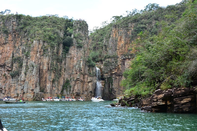 Cachoeira de queda pequena no meio dos Canyons que são grandes pedras nas cores marrons e cinzas com folhagem verde no topo. A cachoeira deságua no grande lago que alguns barcos estão parados. Imagem ilustrando post Airbnb em Escarpas do Lago.