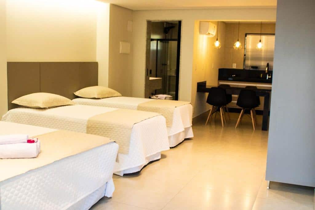 Quarto do ESCARPAS FLATS com três camas de solteiro lado a lado e ao fundo uma porta aberta que dá acesso ao banheiro. Do lado direito uma parte que mostra a cozinha compacta com uma bancada, quatro cadeira, três luminárias no teto, um ar-condicionado e a pia no fundo perto da janela.