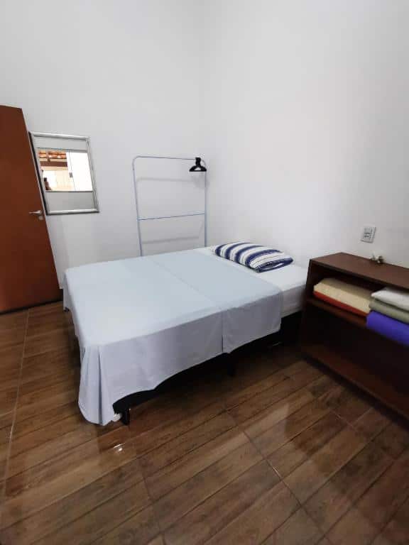 Quarto da Casa do Diogo com uma cama de casal ao centro do quarto, ao lado esquerdo uma cômoda aberta com cobertores guardados e do lado direito uma parede branca com uma arara para pendurar roupas, um espelho na parede e uma porta de madeira.