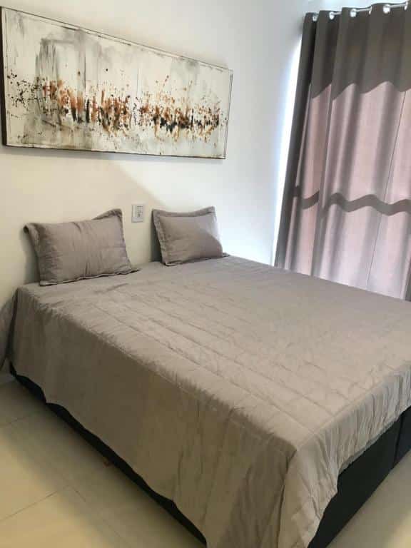 Quarto do Suíte no Mirante em Escarpas com uma cama de casal ao centro com uma colcha e dois travesseiros e na parede um quadro. Do lado direito da cama tem uma cortina cinza.
