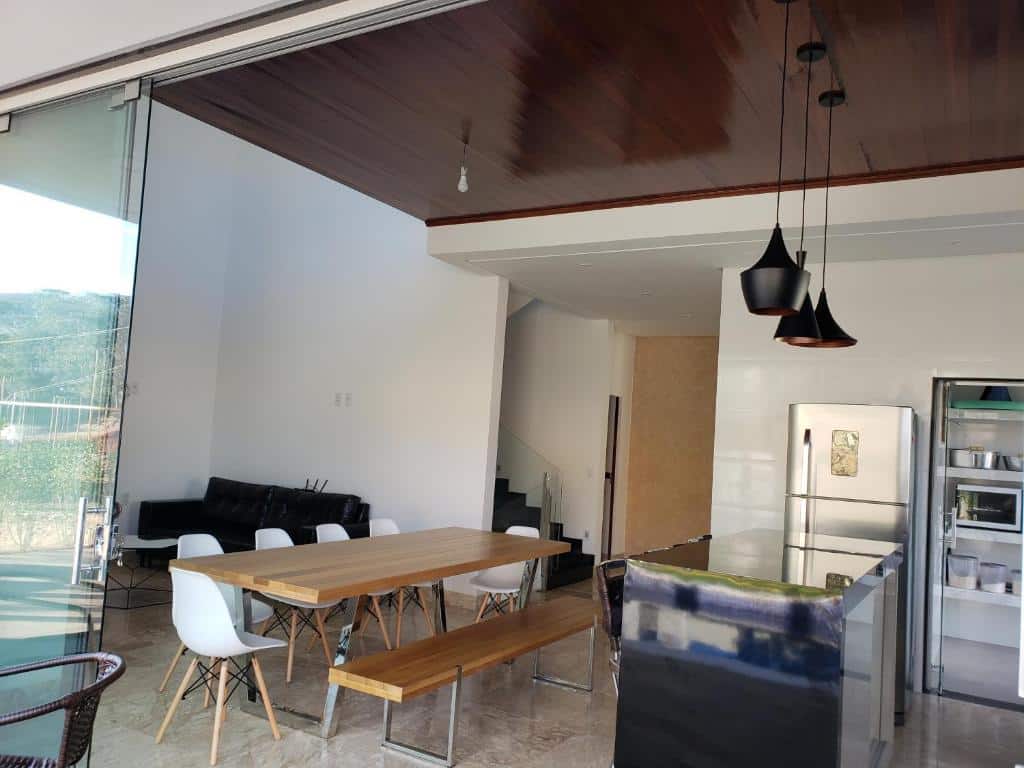 Parte da Casa alto padrão Capitólio Lago Vitória que mostra do lado esquerdo um sofá preto encostado na parede e mais ao centro uma mesa grande de madeira com seis cadeiras e um banco de madeira. Já na parte direita tem um balcão, uma geladeira e do lado um armário.