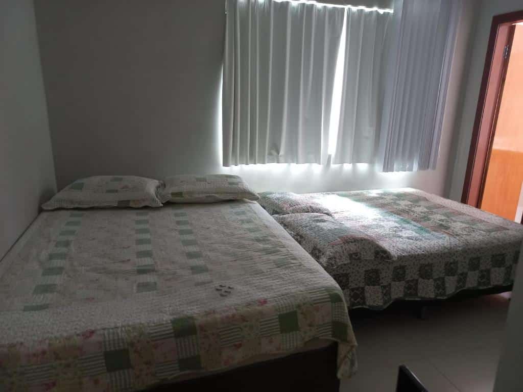 Quarto da Casa Capitolio com duas camas de casal, uma maior encostada na parede esquerda e uma cama de casal menor encostada na parede da janela. Em frente a cama de casal menor tem uma porta de madeira.