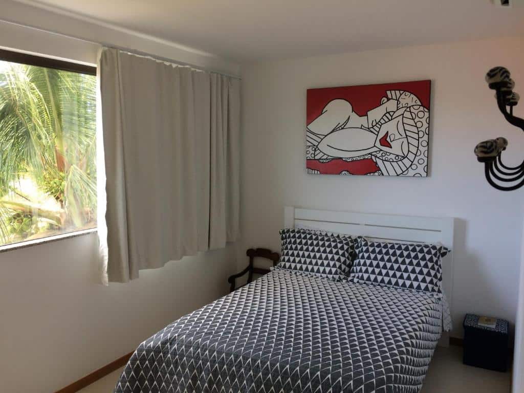 Um quarto pequeno no Salaris Village com uma cama, roupa de cama geométrica, arte na parede acima da cabeceira, janela e cortinas brancas, para representar airbnb em Imbassaí.