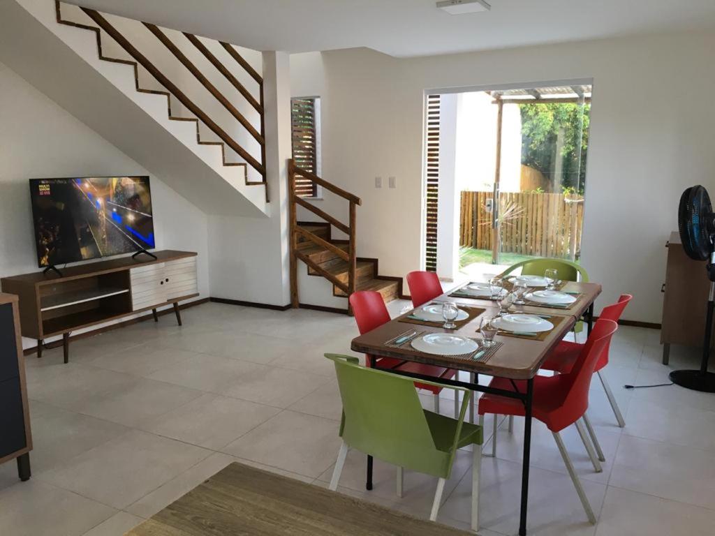 Uma área de jantar com uma mesa posta, cadeiras coloridas, uma TV ligada e uma escada que leva ao andar de cima em uma sala iluminada, na Casa duplex de temporada, para representar airbnb em Imbassaí.