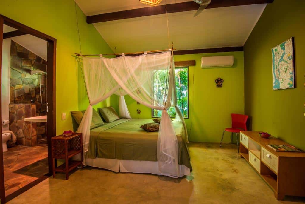 Um quarto na Casa Morada dos Pássaros com paredes verdes, uma cama com dossel branco, uma porta de banheiro aberta, uma janela e móveis simples, para representar airbnb em Imbassaí.