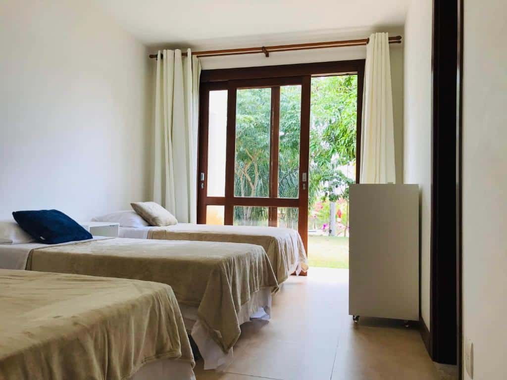Duas camas de solteiro em um quarto bem iluminado com paredes brancas, janela grande, cortinas brancas e vista para a vegetação externa, na Casa Peixinho - Reserva Imbassaí 3 suites, para representar airbnb em Imbassaí.