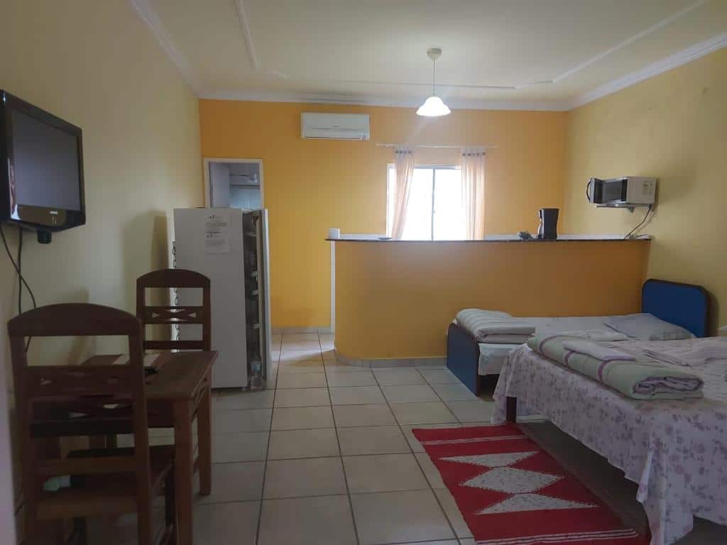 Quarto e cozinha do airbnb Encantu's Flats. No fundo da imagem está a cozinha, que é dividida da sala por uma bancada. Ainda no fundo, no lado direito há um microondas em uma prateleira na parede e no lado esquerdo há uma porta e uma geladeira. Já na frente, no lado esquerdo há uma mesa com cadeiras e uma TV presa a parede e no lado direito há uma cama de casal e uma cama de solteiro lado a lado. Imagem para ilustrar o post airbnb em Itatiaia.