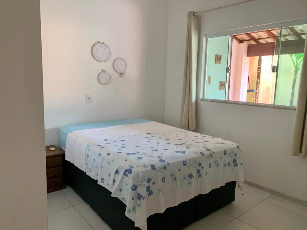 Quarto do airbnb Casa de Férias. No meio do quarto está a cama de casal, no canto esquerdo da cama há uma mesa de cabeceira bem pequena e no lado direito do quarto há uma janela de vidro aberta com cortinas ao lado. Imagem para ilustrar o post airbnb em Itatiaia.