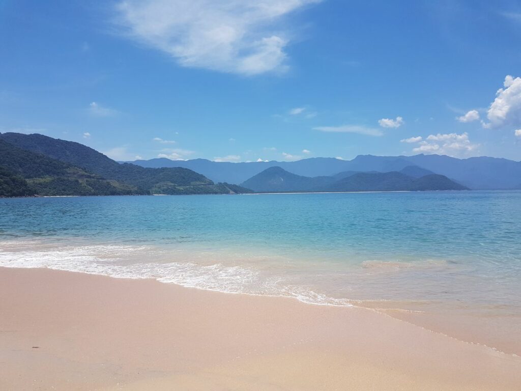 Faixa de areia e mar na Ilha do Prumirim. O mar está calmo e azul. É possível ver algumas montanhas e veegetação ao fundo.