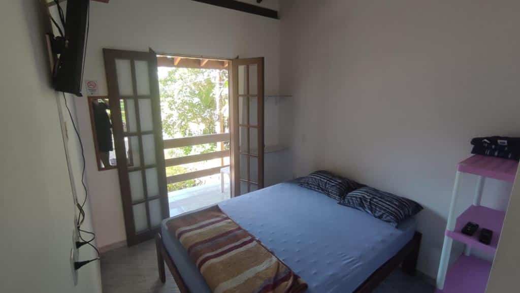 Quarto de um dos Chalés Carioca Prumirim Ubatuba. Uma cama de casal está no meio, ao lado de algumas prateleiras e de frente para uma tv na parede. Ao seu lado esquerdo está a porta para a varanda.