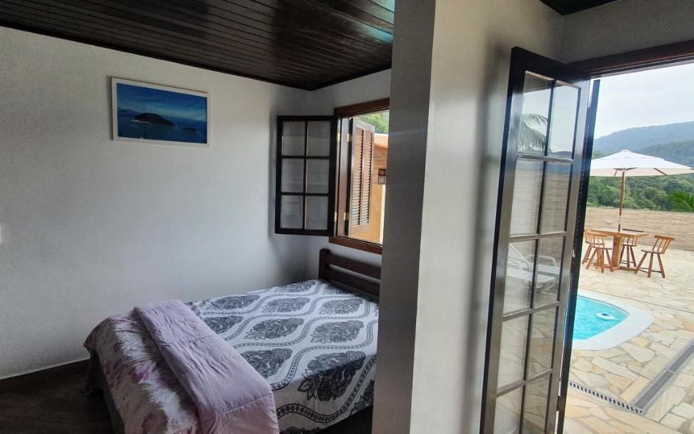 Quarto do Kingston Chalés. Uma cama de casal está ao lado esquerdo da imagem e embaixo de uma janela. Do lado direito uma porta leva até a área da piscina.