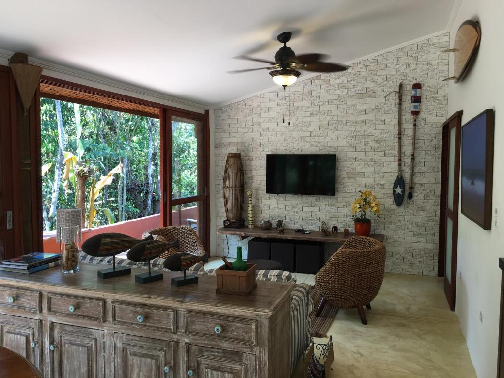 Sala de estar na Beach House Prumirim com cadeiras de vime, uma TV em uma parede de tijolos, decoração de madeira e janelas grandes que mostram a vegetação do lado de fora, para representar airbnb em Prumirim.