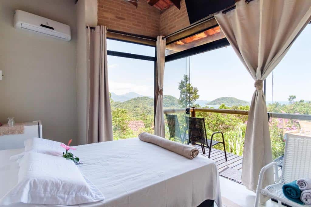 Quarto do Haleiwa chalés e suítes - A Guest House do Prumirim. Uma cama de casal está de frente para uma parede de vidro que mostra toda a paisagem. Uma parte do chão é de madeira e forma a varanda com duas cadeiras lado a lado.
