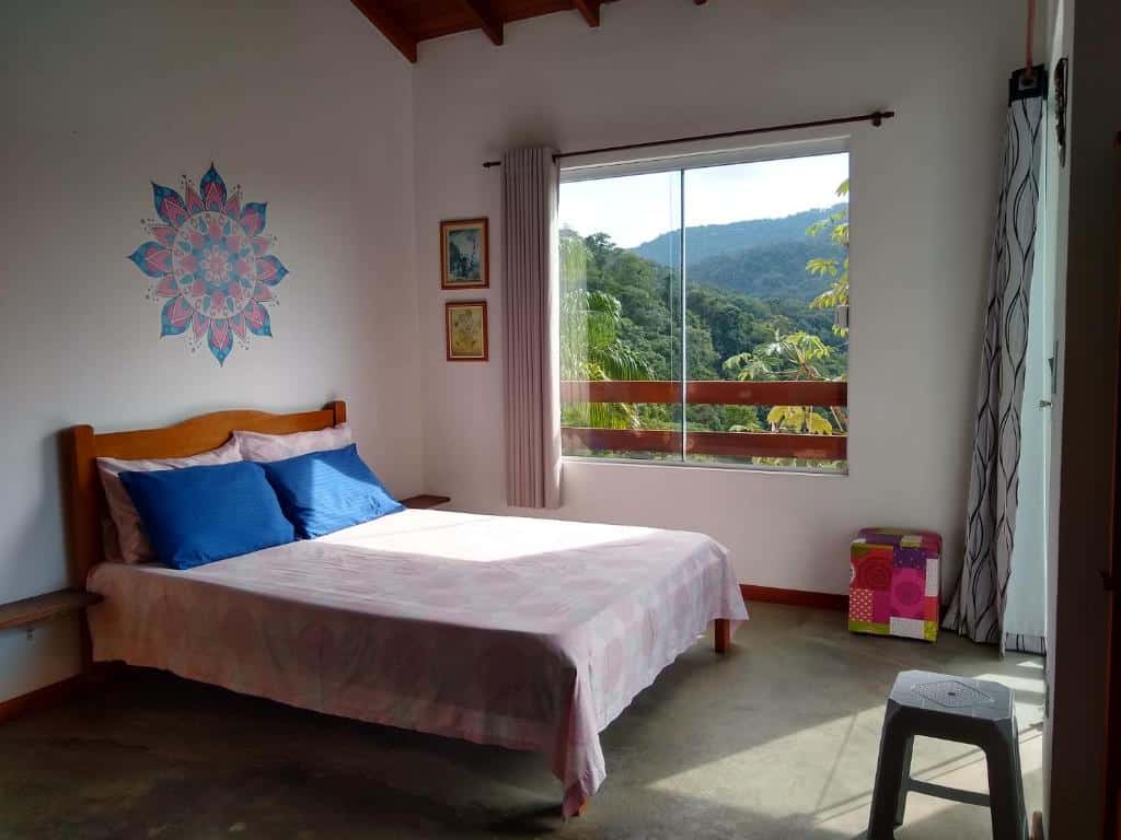 Quarto do La Casa Rozada Prumirim. Uma cama de casal está ao lado de uma janela, que mostra a paisagem com árvores do lado de fora. Perto da cama há também dois bancos.