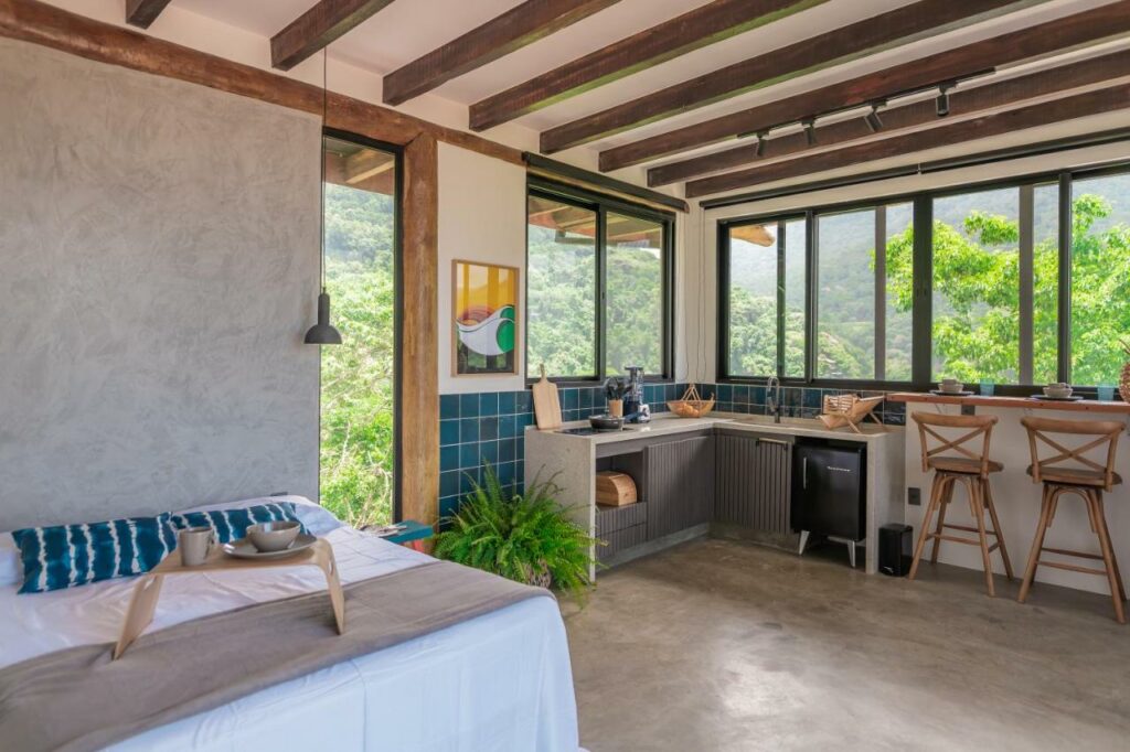 Quarto e cozinha da Casa Ocean. Ao lado esquerdo está a cama de casal. Do lado direito está um balcão, fogão, uma minigeladeira, armários e alguns utensílios. Todo o cômodo possui janelas de vidro e é rodeado de árvores.