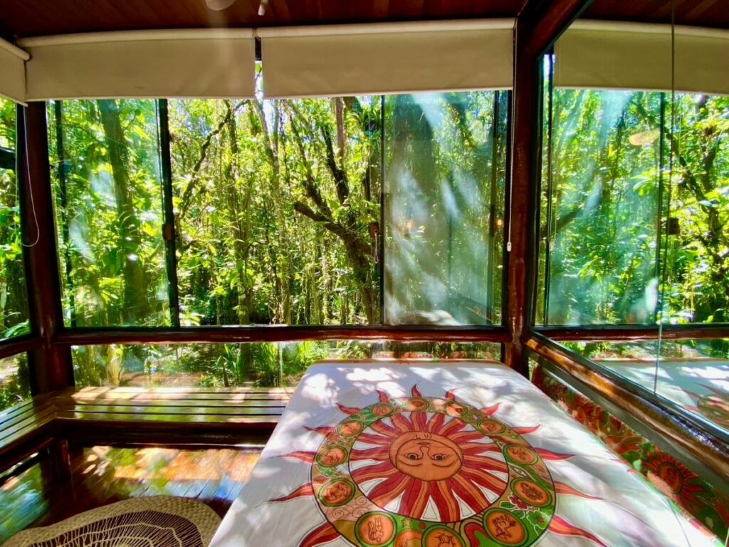 Quarto do Housing 31 durante o dia com cama de casal do lado direito da imagem as paredes são de vidro que tem vista para a natureza. Representa airbnb em São Sebastião.