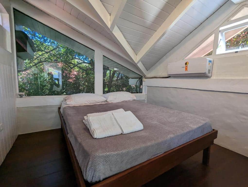 Quarto da Linda Casa Praia de Pauba em condomínio com uma cama com roupa de cama cinza e toalhas dobradas fica sob tetos brancos inclinados, ao lado de grandes janelas que mostram a vegetação do lado de fora. Representa airbnb em São Sebastião.