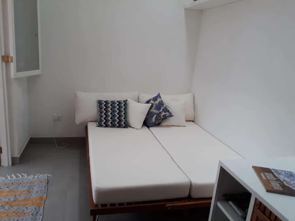 Um pequeno quarto do lindo chalé em Boiçucanga Condomínio com um sofá-cama branco, travesseiros estampados e sólidos, um tapete e uma pequena prateleira branca com livros. Representa airbnb em São Sebastião.