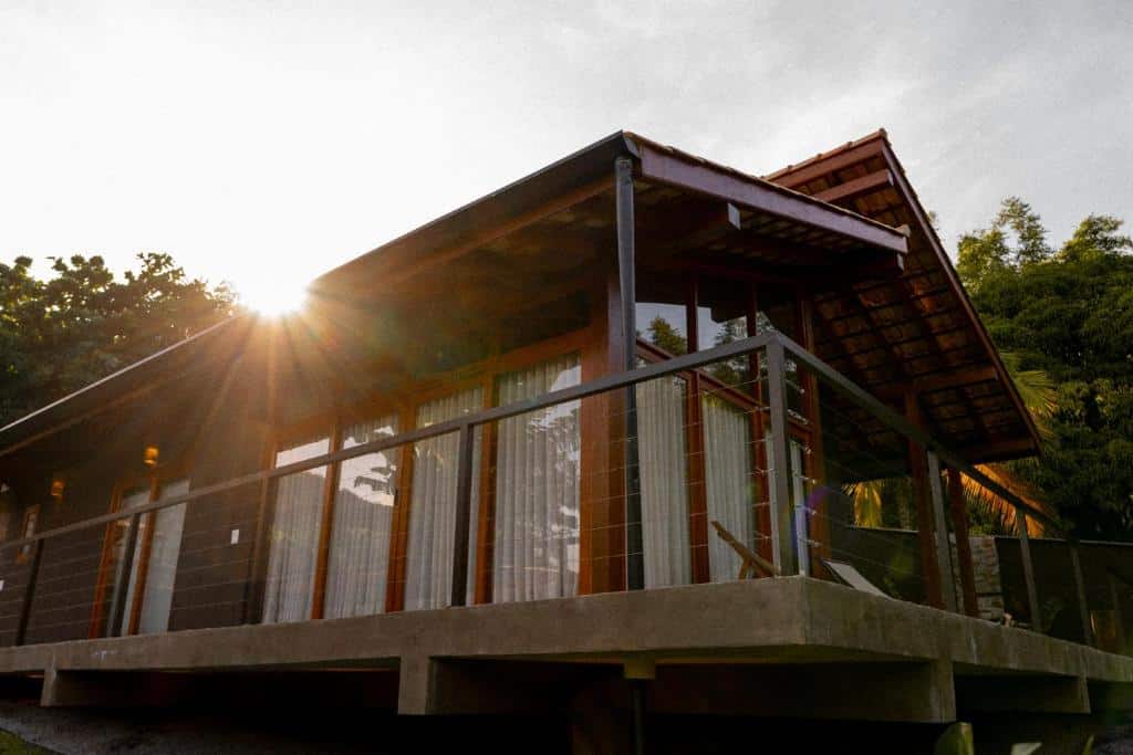 Casa térrea Nai com grandes janelas e um deck envolvente, cercada por vegetação, com a luz do sol brilhando por trás.