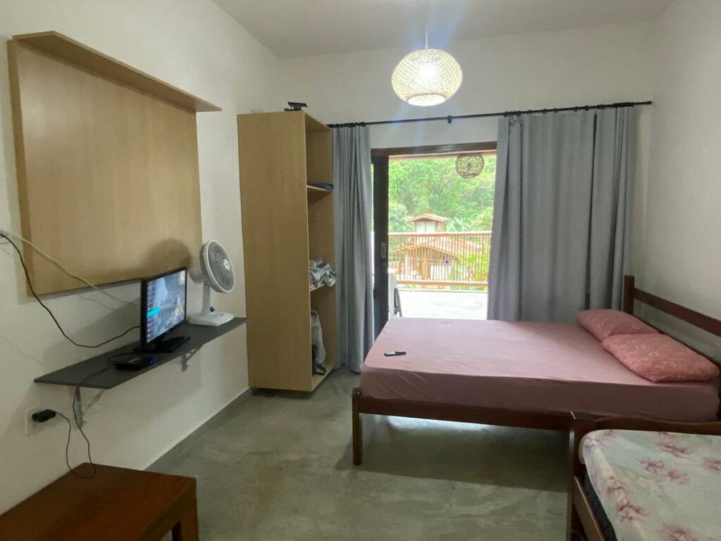 Quarto do Loft encantador Soul Happy. Uma cama de casal está no meio, entre a porta da varanda e um sofá-dama. Em frente a ela está um armário, uma tv e um ventilador, para representar airbnb na Praia Vermelha do Centro.