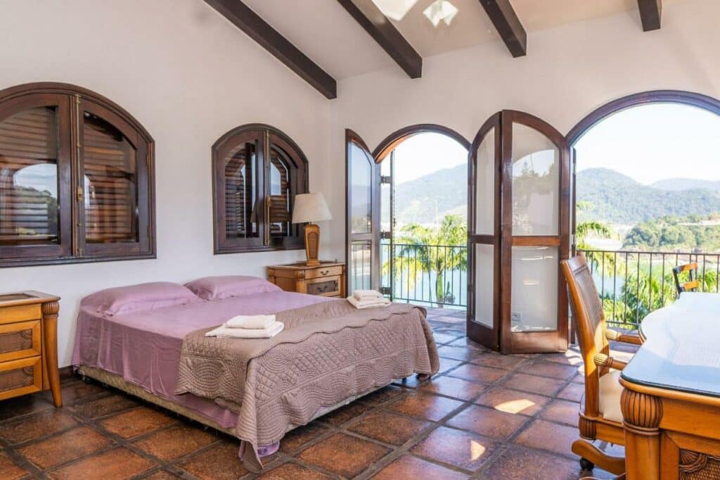 Um dos quartos da Casa luxo, com 9 quartos vista mar Ubatuba. A cama de casal está no meio, entre duas mesas de cabeceira e de frente para uma mesa de trabalho na outra parede. Ao lado da cama estão as portas para a varanda que possui vista para o mar. Ele faz parte da lista de airbnb na Praia Vermelha do Centro.