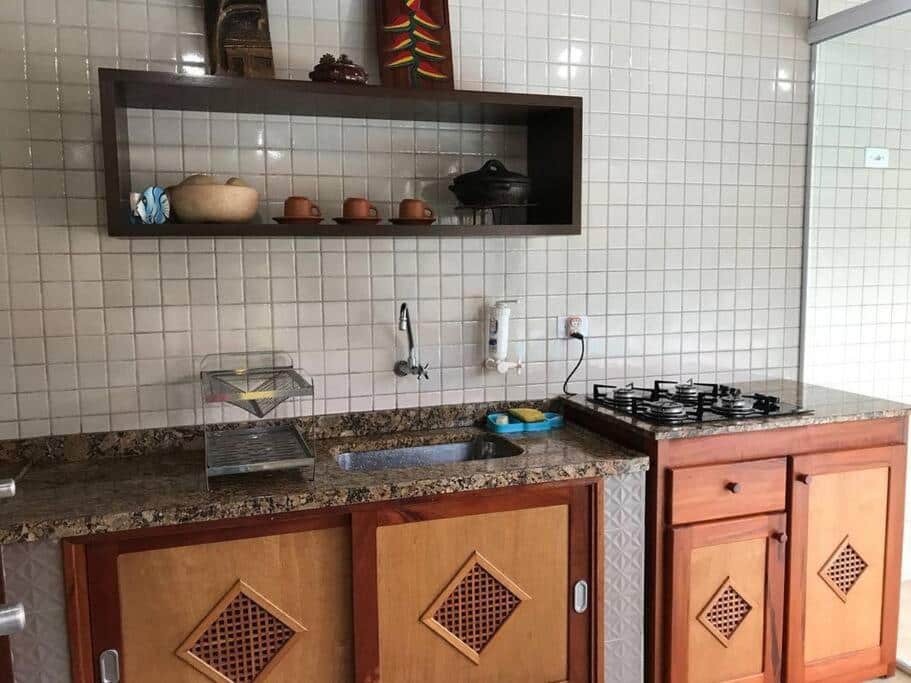 Cozinha da Casa dentro cond. Tenório. Uma pia e um fogão estão lado a lado, abaixo deles um armário. Na parede acima da pia há uma prateleira com xícaras e outros objetos.