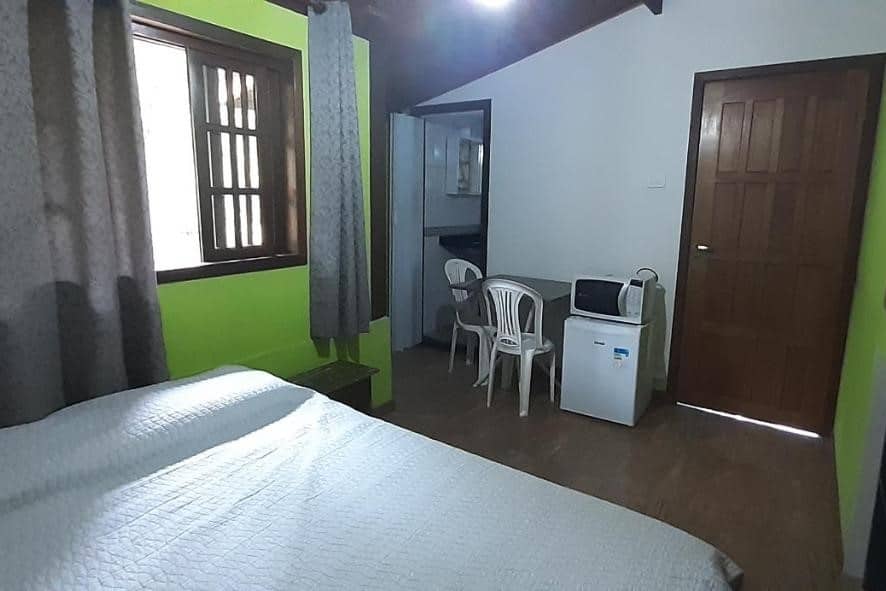 Quarto da Suíte Som do Mar da Vermelha do Centro. Uma cama de casal está de frente para uma mesa de dois lugares, um frigobar e um micro-ondas, todos na parede oposta. Ao lado deles está a porta para oo banheiro e a porta de entrada, airbnb na Praia Vermelha do Centro.