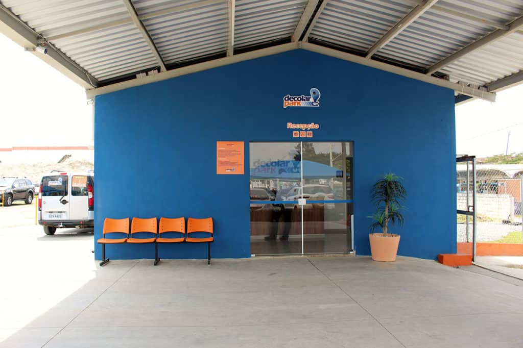 Recepção do Decolar Park com uma porta de vidro, algumas cadeiras do lado de fora e uma cobertura para proteger do sol e chuva