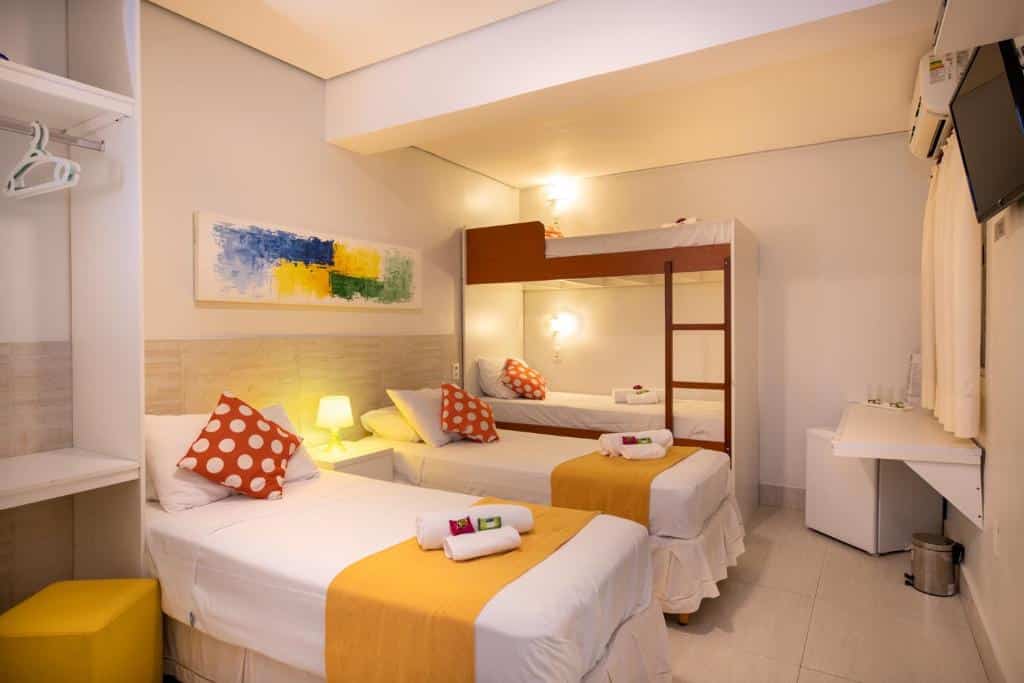 Um quarto bem iluminado com duas camas de solteiro, um beliche, detalhes em amarelo e uma TV na parede. Esse é o Brazilodge All Suites Hostel.