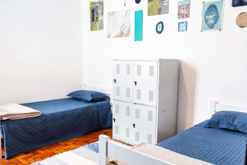 Quarto do Neighbor.Hub com duas camas de solteiro com roupa de cama azul, um armário de metal no meio e obras de arte em uma parede branca acima das camas.