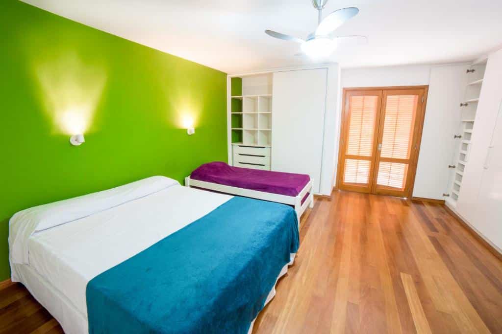 Quarto do Madá Hostel com duas camas, parede com detalhes em verde, prateleiras embutidas, piso de madeira e portas duplas com persianas.