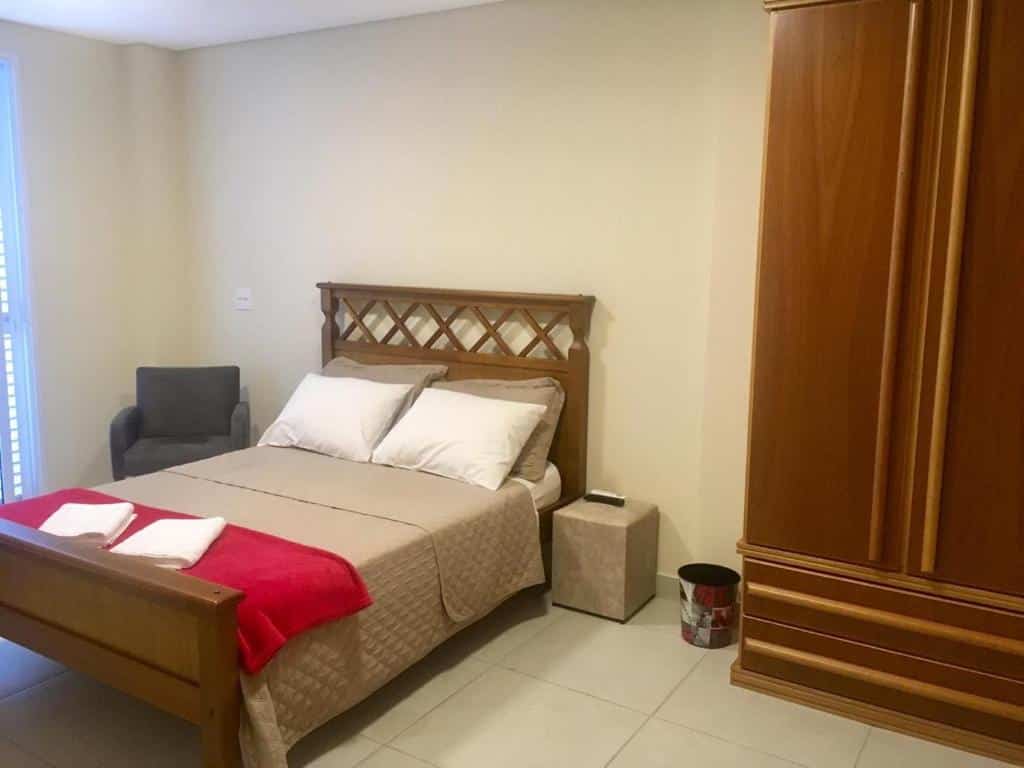 Uma cama de casal bem arrumada no Tapera Hostel com roupa de cama bege, dois travesseiros, um cobertor vermelho, uma cadeira cinza e um guarda-roupa de madeira.