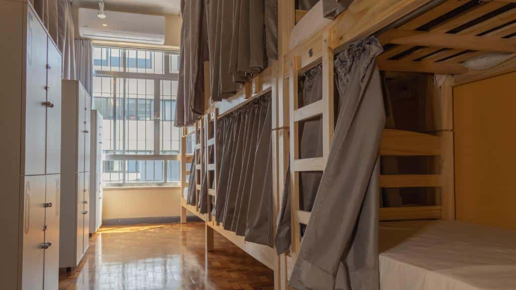 Quarto com beliches de madeira, cada cama tem cortinas de privacidade; armários e uma grande janela permitem a entrada de luz natural. Esse é o Hostel Cuarto.