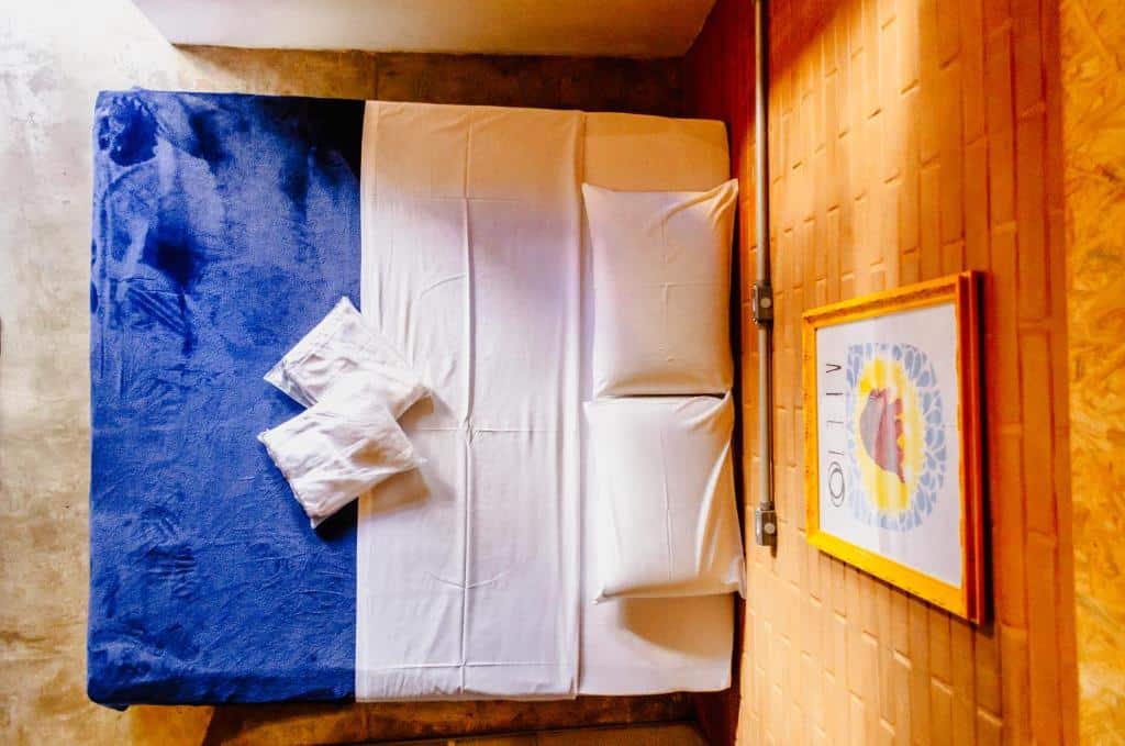 Uma cama bem arrumada no O de Casa Hostel Bar com lençóis brancos, dois travesseiros e um cobertor azul, ao lado de uma parede de madeira com uma foto emoldurada.