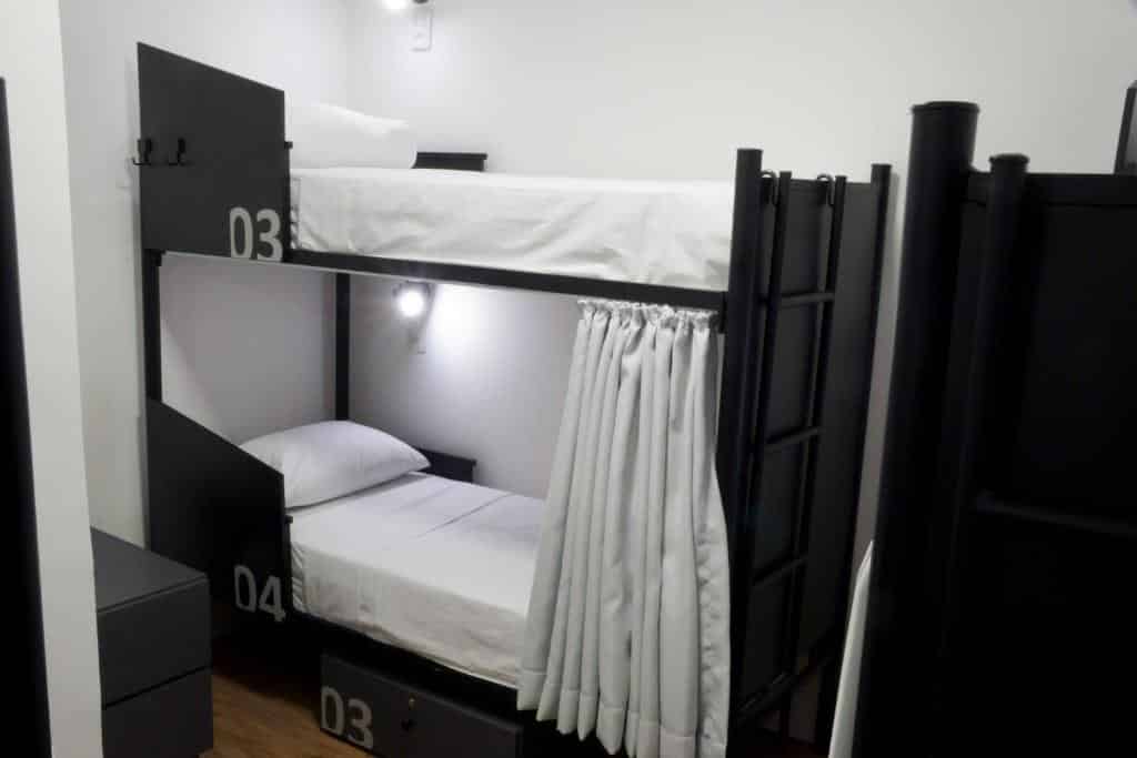 Um quarto moderno do Hostel More SP com beliches de metal preto, numerados como 03 e 04, ambos com roupa de cama branca e cortinas de privacidade.