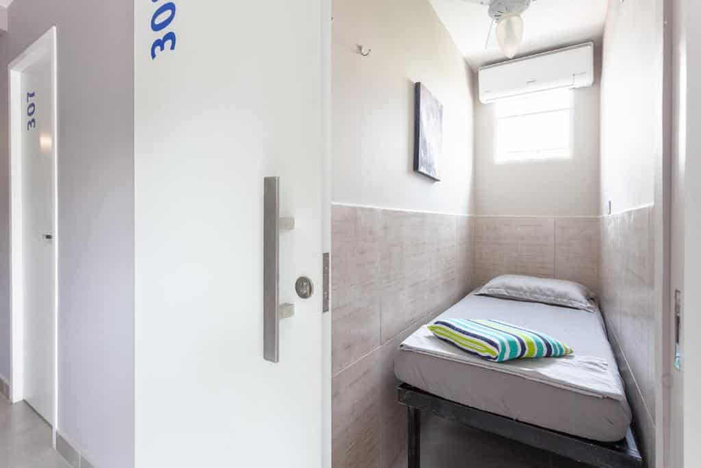 Quarto pequeno e estreito do São Paulo Hostel Club com uma cama de solteiro, roupa de cama cinza, travesseiro listrado, janela e ar-condicionado acima da cama.
