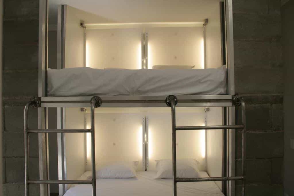 Um beliche com estrutura de metal, com roupa de cama e travesseiros brancos, iluminado por luzes embutidas em uma parede simples. Esse é o Viva Hostel Design.