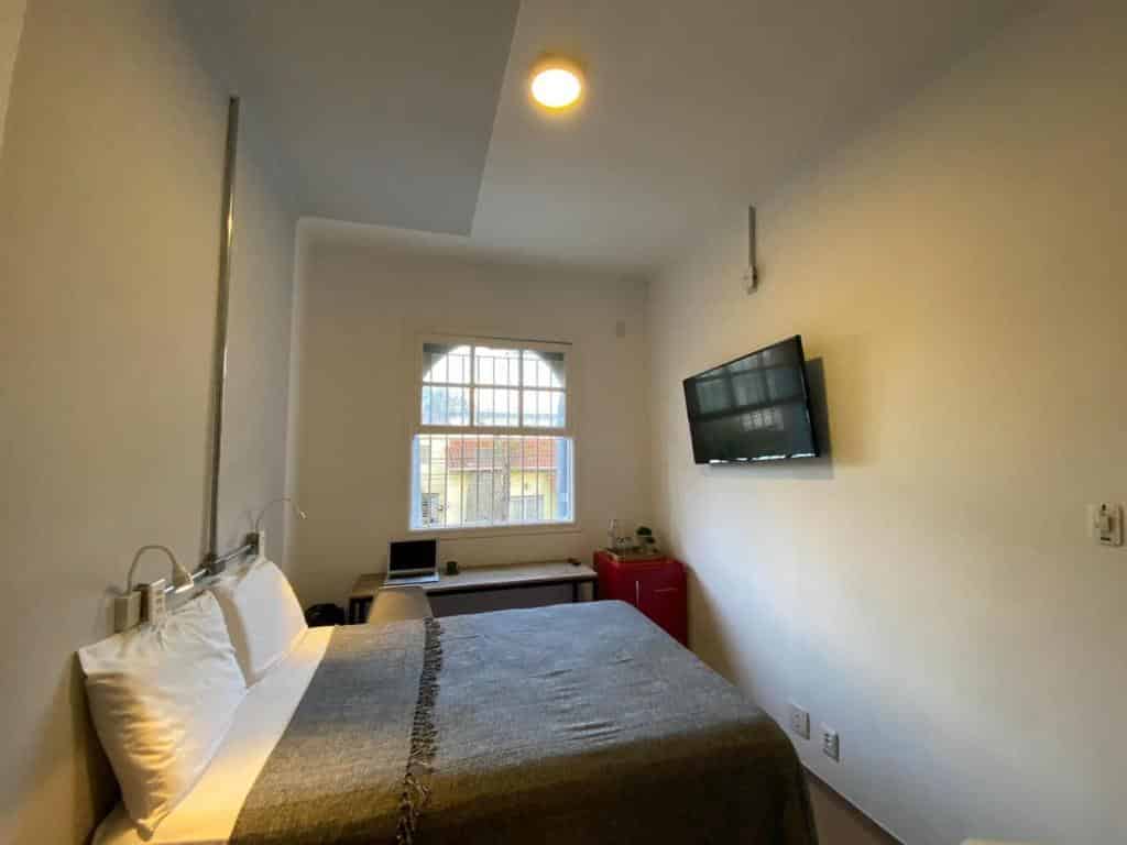 Quarto pequeno do Angatu Hostel com uma cama feita, TV na parede, janela, escrivaninha com laptop e um frigobar vermelho.