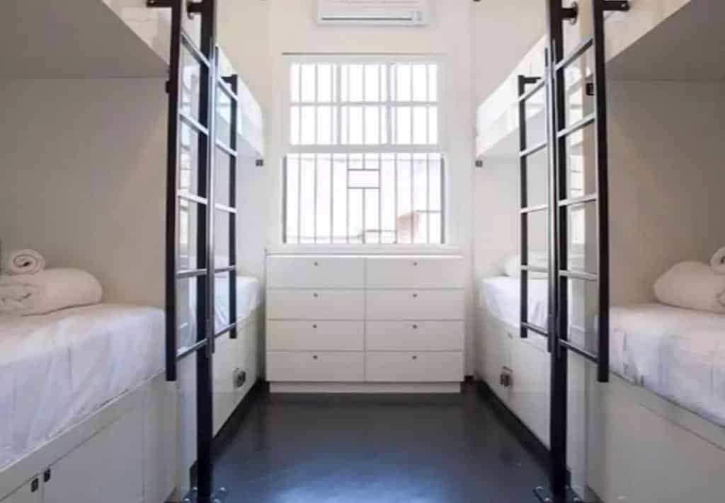 Um quarto do Hostel Brasil Boutique com quatro beliches, escadas, roupa de cama branca, uma janela com grades e uma cômoda branca embaixo da janela.