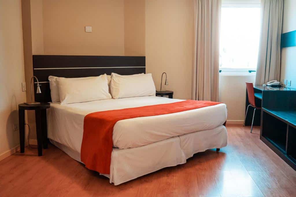 quarto do Mérit San Telmo com cama de casal na diagonal com mesinha e luminária de ambos os lados, há uma janela ampla com cortinas do outro lado, em frente há uma mesinha com cadeira 