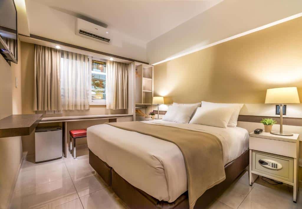 quarto do Up Recoleta Hotel com cama de casal, mesinha e luminária em ambos os lados, escrivaninha comprida com cadeira abaixo da janela larga com cortinas curtas, em frente há uma tv