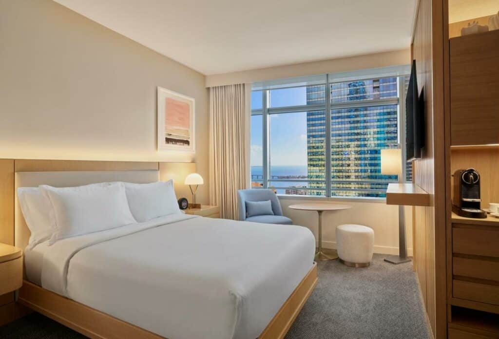 Imagem do quarto do Hotel AKA Brickell com cama de casal do lado esquerdo da imagem no centro do quarto com uma comoda em cada lado com luminária do lado esquerdo da cama uma poltrona com uma mesa redonda e um banco redondo ao fundo uma janela com vista para a cidade em frente a cama uma TV presa na parede. Representa hotéis 5 Estrelas em Miami.