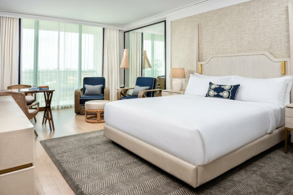 Imagem do quarto do The Ritz-Carlton Coconut Grove, Miami com cama de casal do lado direito da imagem, do lado direito uma cômoda com luminária, ainda do lado direito duas poltronas com um banco estofado no centro. Representa hotéis 5 Estrelas em Miami.