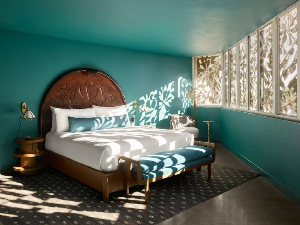 Imagem do Mayfair House Hotel & Garden com cama de casal do lado esquerdo da imagem no centro do quarto com uma cômoda em cada lado da cama, no pé da cama um banco estofado e do lado esquerdo da cama uma poltrona com uma luminária do lado esquerdo. Representa hotéis 5 Estrelas em Miami.