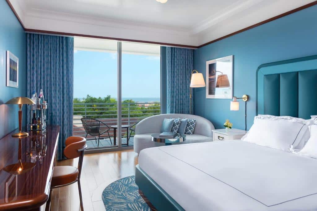 Quarto do Mr. C Miami – Coconut Grove com cama de casal do lado direito da imagem a frente, em frente a cama uma mesa com cadeira e do lado direito da cama uma cômoda e um sofá com mesa de centro e ao fundo uma porta de vidro com acesso a varanda. Representa hotéis 5 Estrelas em Miami.