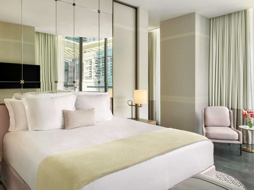 Quarto do SLS LUX Brickell com cama de casal do lado esquerdo da imagem do lado esquerdo da cama uma cômoda com luminária e no canto uma poltrona. 